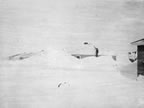 Thumbnail photo of man standing on tall snowdrift touching the roof of a house.