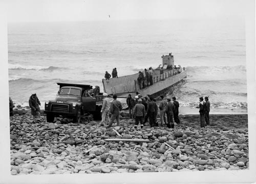 Photo of people on rocky beach driving a truck off of a landing craft.