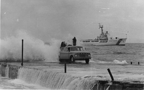 Photo of water washing over truck, with a Coast Guard ship in the background.