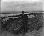 Thumbnail photo of a man in a suit and hat standing on a rocky beach.