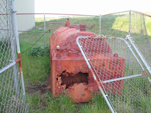 Photo of a rusty machine behind a half-collapsed chain link fence, the School AST.
