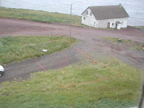 Photo of a rectangular area marked by stakes and tape, with a weathered white building in the background.