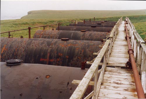 Photo of a row of rusted tanks with wooden walkway at the Abandoned Diesel Tank Farm, prior to remediation.