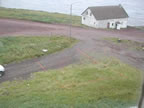 Thumbnail photo of a rectangular area marked by stakes and tape, with a weathered white building in the background.