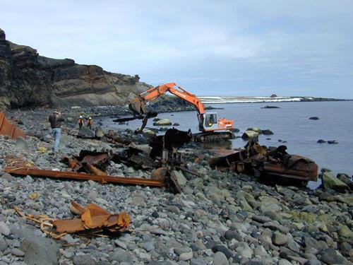 Photo of an excavator removing metal debris from a rocky shore.