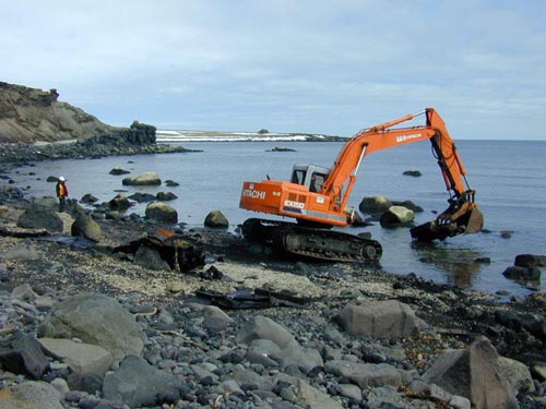 Photo of an excavator removing metal debris from a rocky shore.