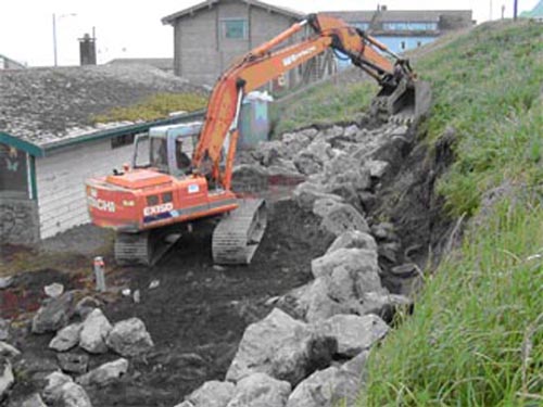 Photo of an excavator placing boulder-sized rocks at the base of a hill.