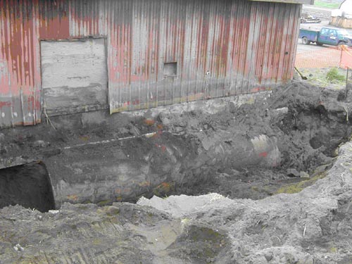 Photo of a deep trench at the base of a weathered red building.