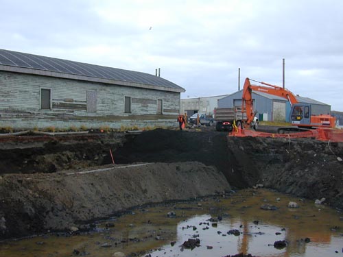 Photo of a deep water-filled ditch with a weathered building and an excavator in the background.