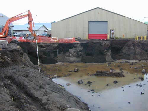 Photo of a deep water-filled ditch with a weathered building and excavator in the background.