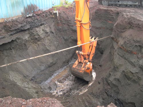 Photo of an excavator digging hole at the base of a blue building.