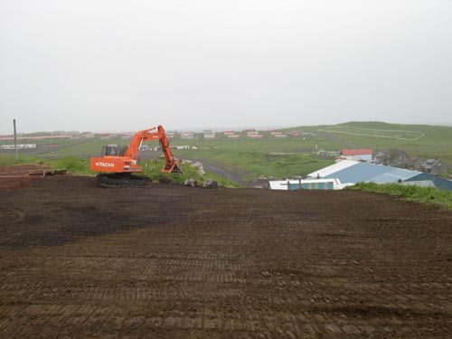 Photo of a large open dirt area with an excavator and buildings in the distance.