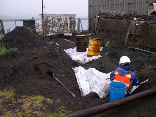Photo of a large pile of dirt with shovel, two drums on white plastic, and person in hard hat with a blue bucket.