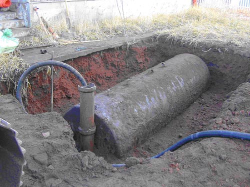 Photo of a hole in the ground with a large partially-buried cylinder with attached hose.