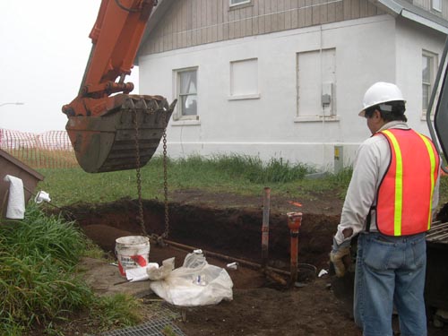 Photo of an excavator with chain attached to a cylinder in ground. Person with hard hat is in the foreground.