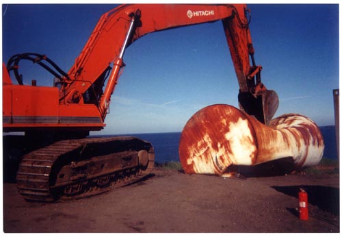 Photo of an excavator crushing a large rusted drum.