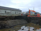 Thumbnail photo of deep water-filled ditch with weathered building and excavator in the background.