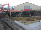 Thumbnail photo of a deep water-filled ditch with a weathered building and excavator in the background.