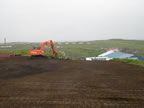 Thumbnail photo of large open dirt area with excavator and buildings in the distance.
