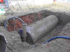 Thumbnail photo of hole in the ground with large partially-buried cylinder with attached hose.