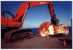 Thumbnail photo of an excavator crushing a large rusted drum.