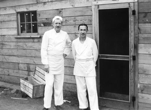 Photo of two men in white outside of wooden building.