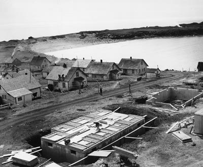 Photo of a collection of small houses on one side of a dirt road with a building under construction on the other side of the road.